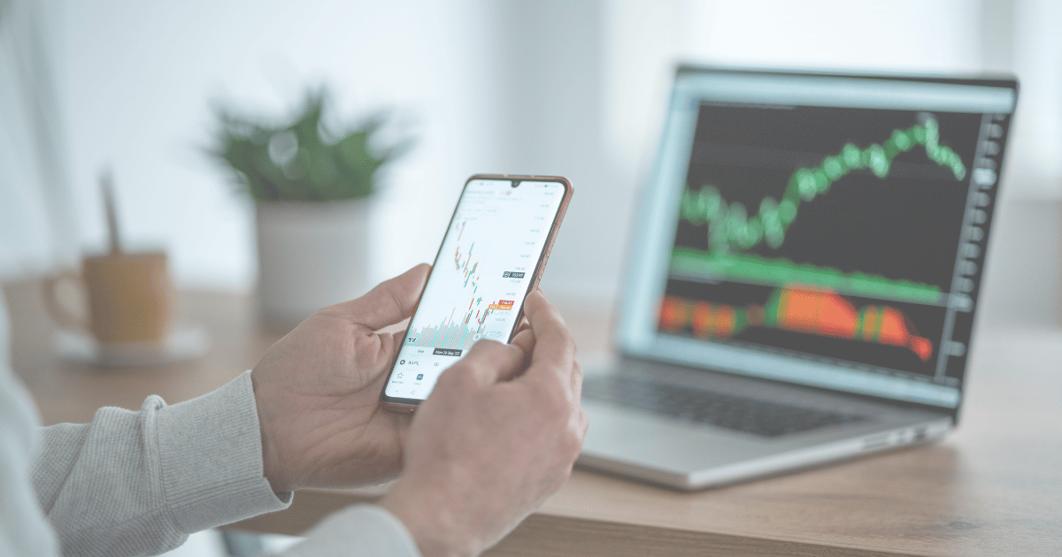person holding phone in front of laptop with stock graph on both screens